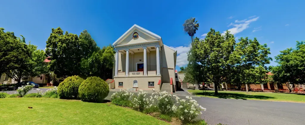 The Japie Krige Hall seen down the school driveway: a cream building with tall white columns, PAUL ROOS GIMNASIUM across the pediment and a JAPIE KRIGE SAAL / HALL sign below it.
